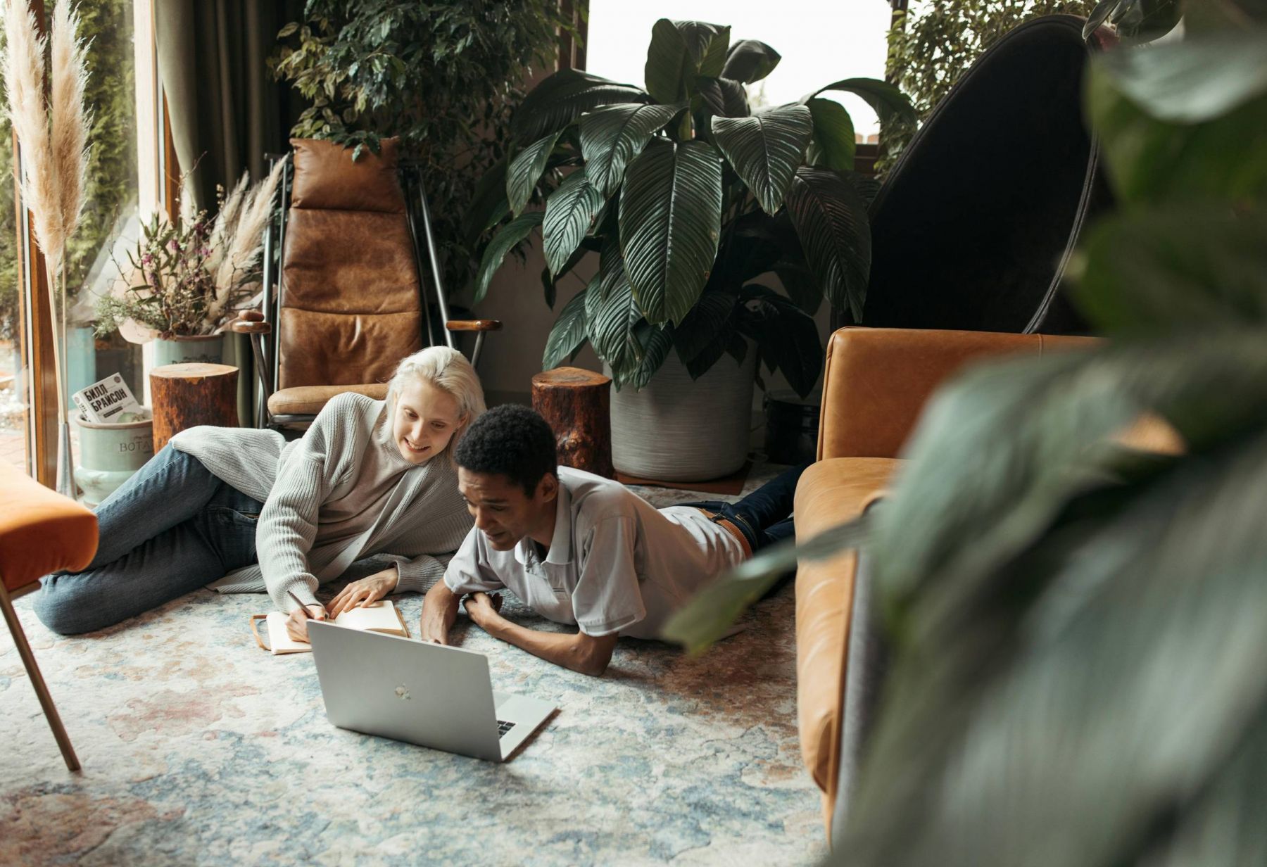 Ever College Station student living apartments Two people lying on the floor, smiling and looking at a laptop in a cozy, plant-filled living room.