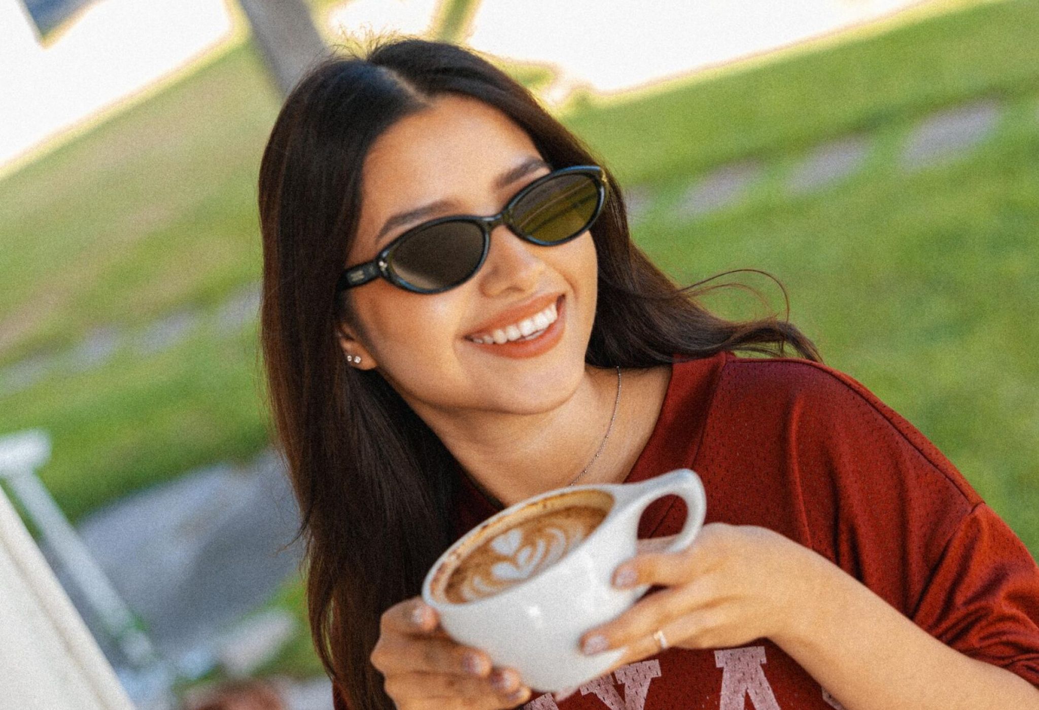 Ever College Station student living apartments A smiling woman wearing sunglasses and a Texas A&M shirt holds a cup of latte art coffee.