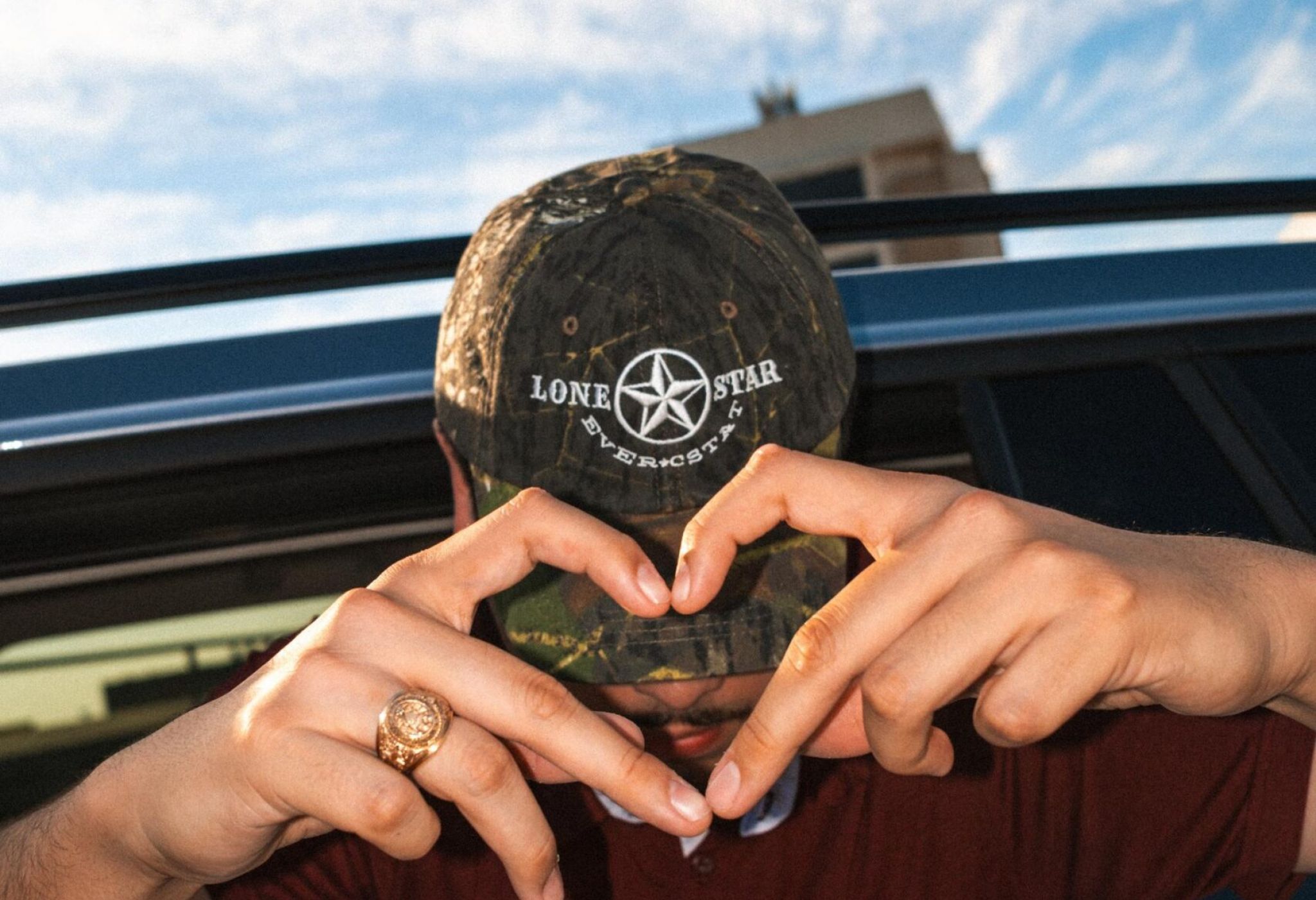 Ever College Station student living apartments A person in a camouflage cap and maroon shirt making a heart shape with their hands while leaning out of a car window.
