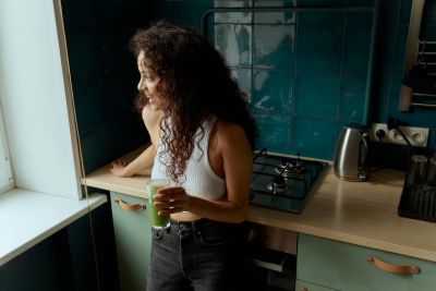 Ever College Station student living apartments Woman with curly hair holding a green drink, standing in a kitchen and looking out the window.