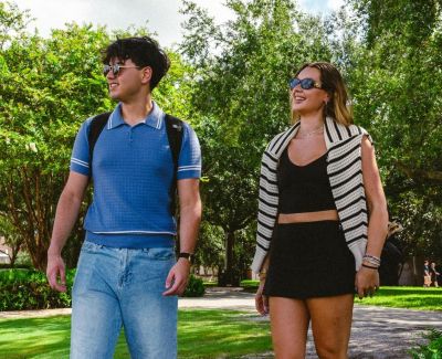Ever College Station student living apartments Two people wearing sunglasses walk together outside on a sunny day, surrounded by green trees and grass.