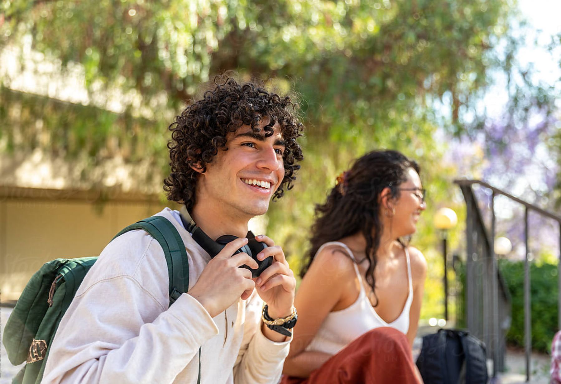 EVER College Station Smiling young man with headphones and backpack sits outdoors with a woman in the background on a sunny day.
