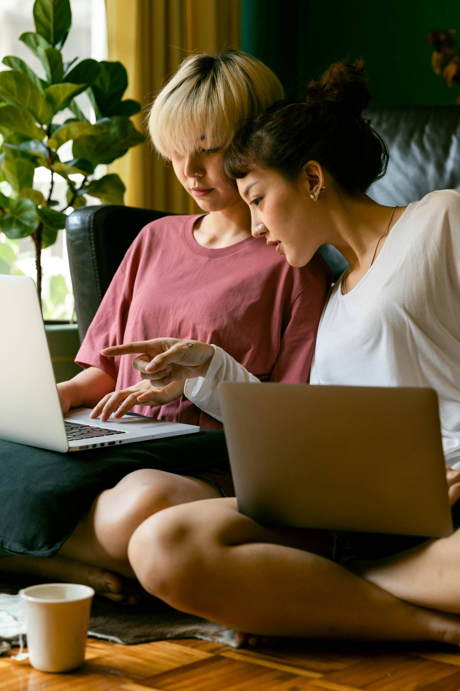 Ever College Station student living apartments Two women sit on the floor using laptops, one pointing at the other's screen, near a window and plants.