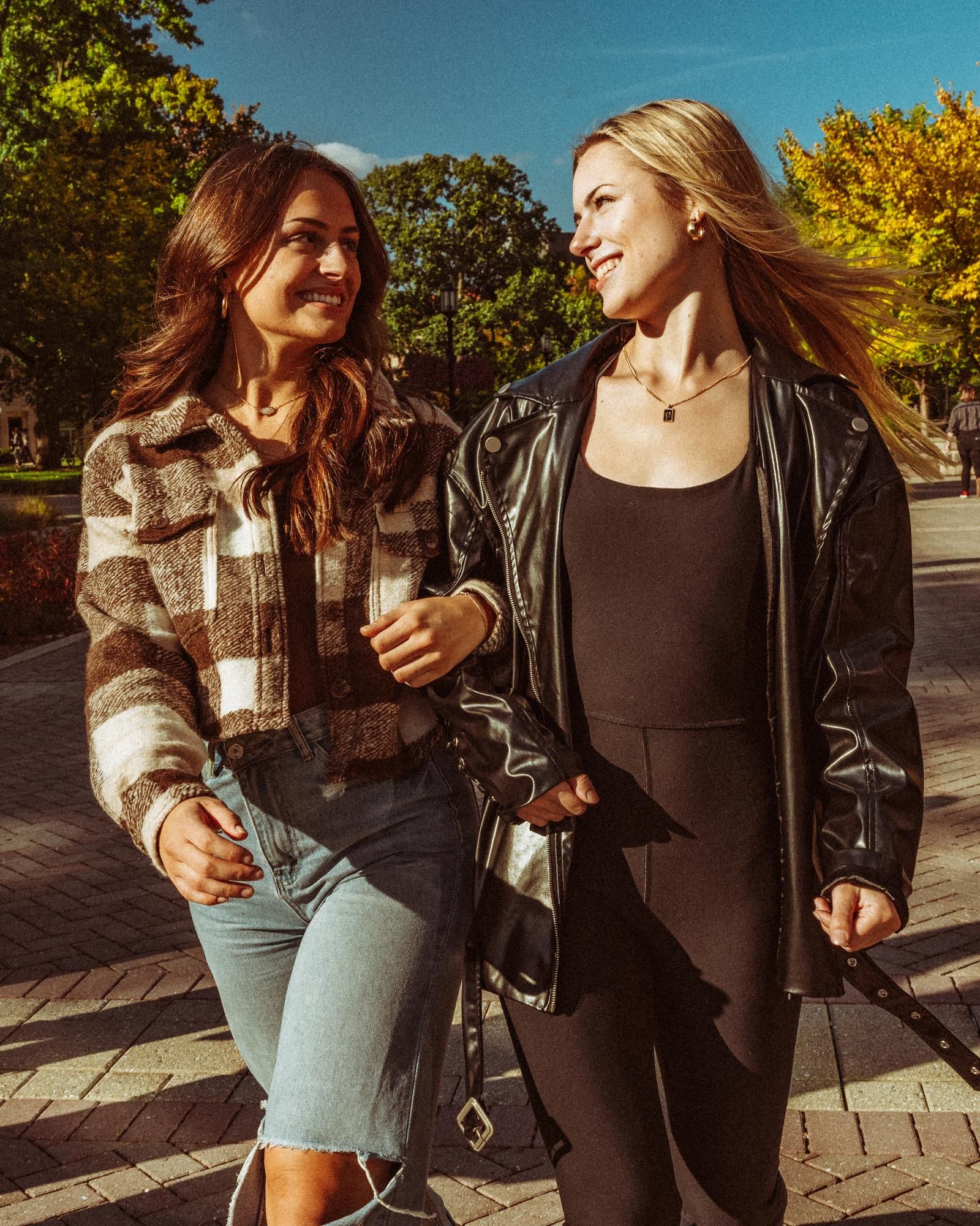 Ever College Station student living apartments Two women walking outdoors on a sunny day, smiling at each other, arm in arm on a paved path.
