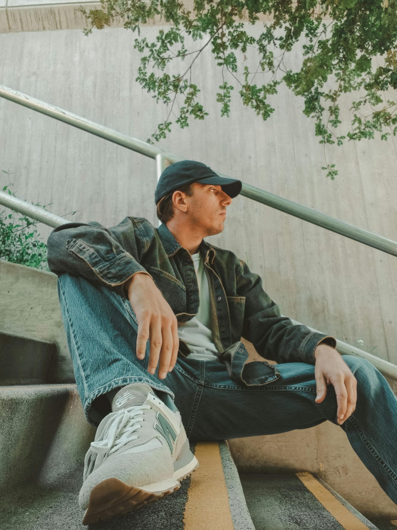 EVER College Station Man in casual clothes and cap sitting on stairs outdoors, looking to the side under leafy branches.